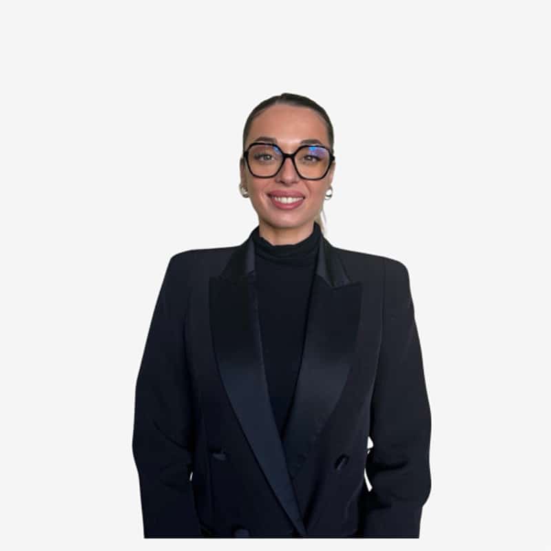 Femme d'affaires souriante en blazer noir et lunettes Femme souriante, brune aux cheveux tirés, portant des lunettes noires oversize et un blazer noir à revers satiné sur un col roulé noir.